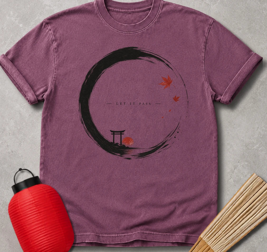 A mauve purple short-sleeve t-shirt with a black circular brushstroke graphic, a small torii gate, and red maple leaves with the text 'LET IT PASS'. The shirt is styled in a flat-lay on a gray textured background with a red paper lantern and wooden chopsticks.